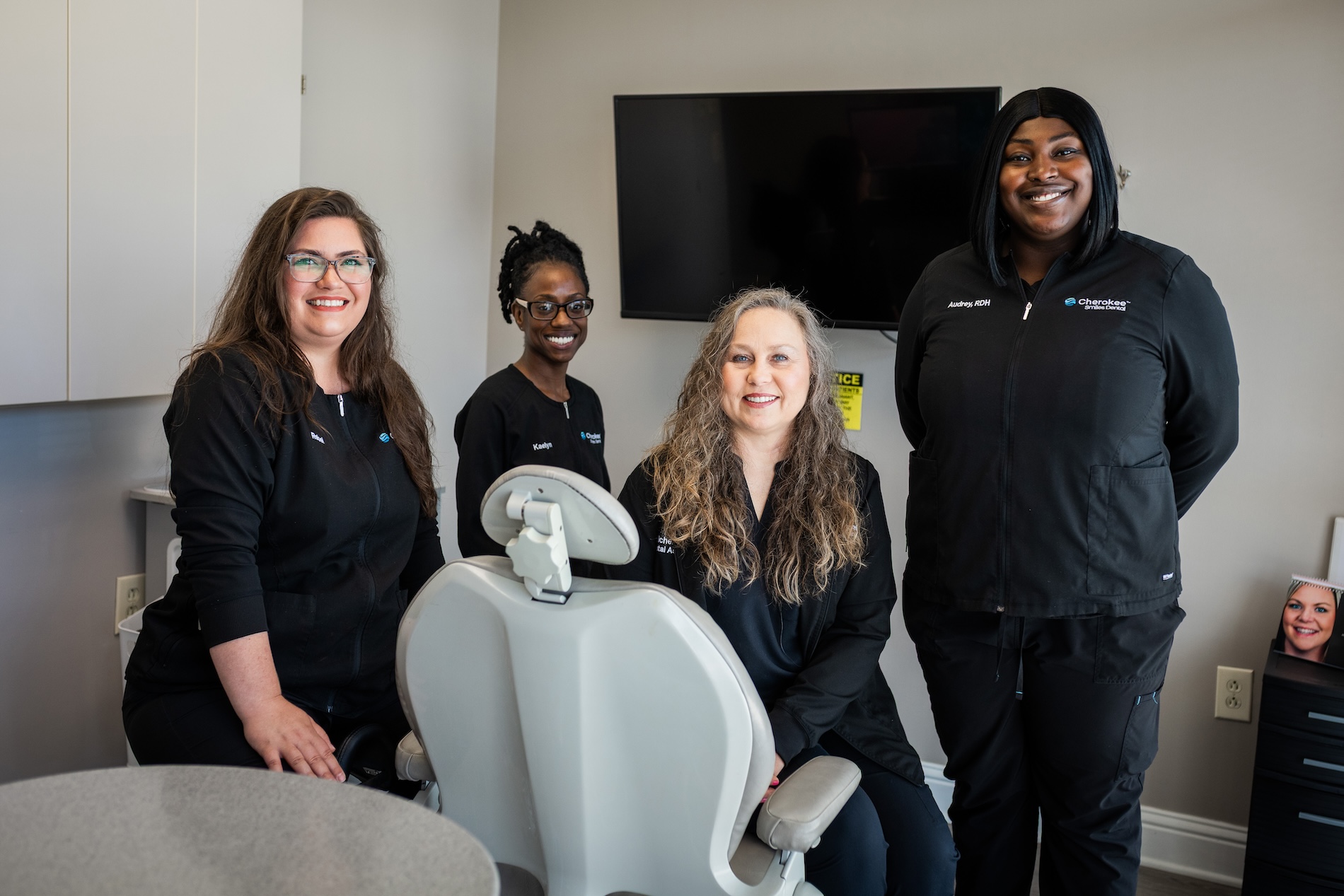 The image shows four individuals posing with a dental chair in an office setting, with a sign indicating Dental Hygiene.