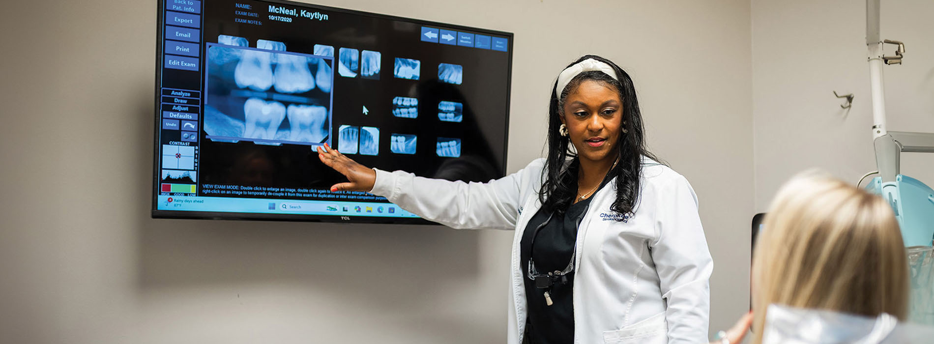 A dental professional stands in front of an X-ray machine, gesturing towards a large screen displaying dental images.