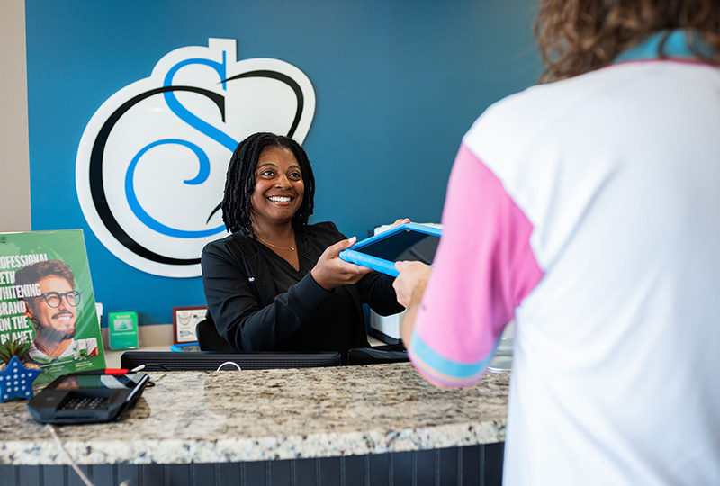 The image shows an indoor scene with a person standing at a counter, holding a blue object that could be a pen or stylus, interacting with another individual who appears to be behind the counter. The setting suggests a retail or service establishment, as indicated by the branded sign on the wall and the presence of promotional materials in the background.