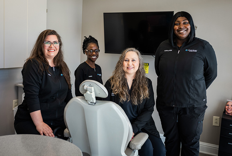 The image shows four individuals standing around a dental chair in a dental office setting, with one person seated on the chair and two others standing behind it. They are all wearing face masks, and there s a TV screen displaying a smiling face on the wall behind them.