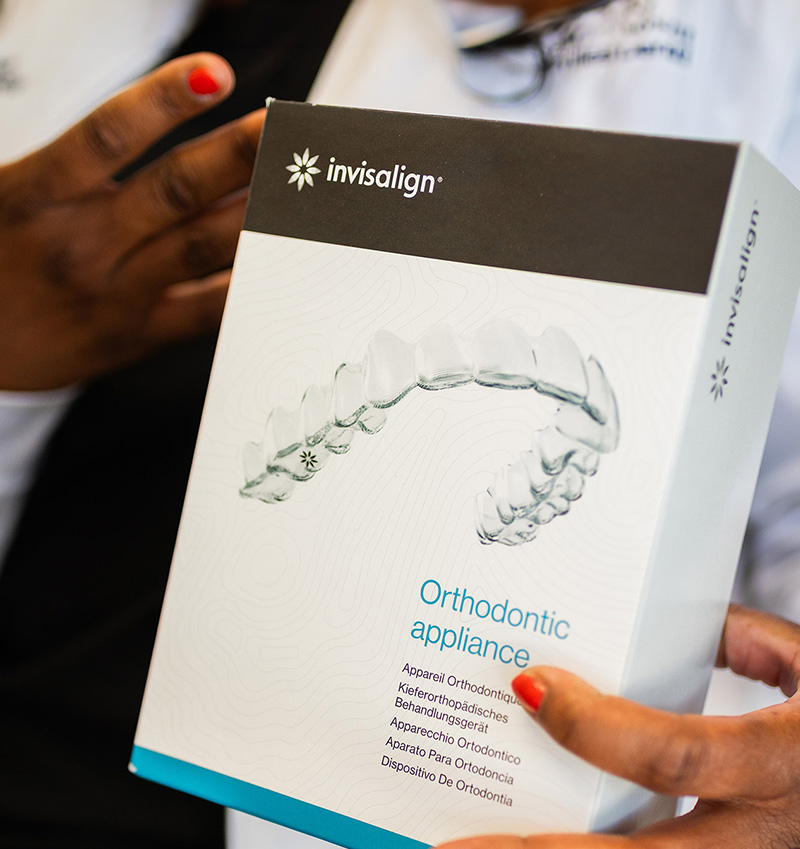 The image shows a person holding a boxed orthodontic appliance with an open lid, revealing the clear aligners inside, against a blurred background.