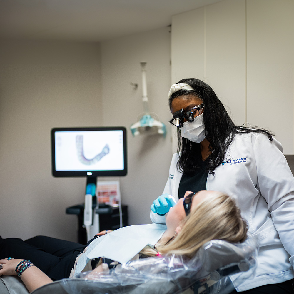 The image shows a dental hygiene professional wearing protective eyewear and a face mask while sitting at a dental chair with a patient receiving treatment, likely a cleaning or procedure, with both of them wearing face masks and following safety protocols.