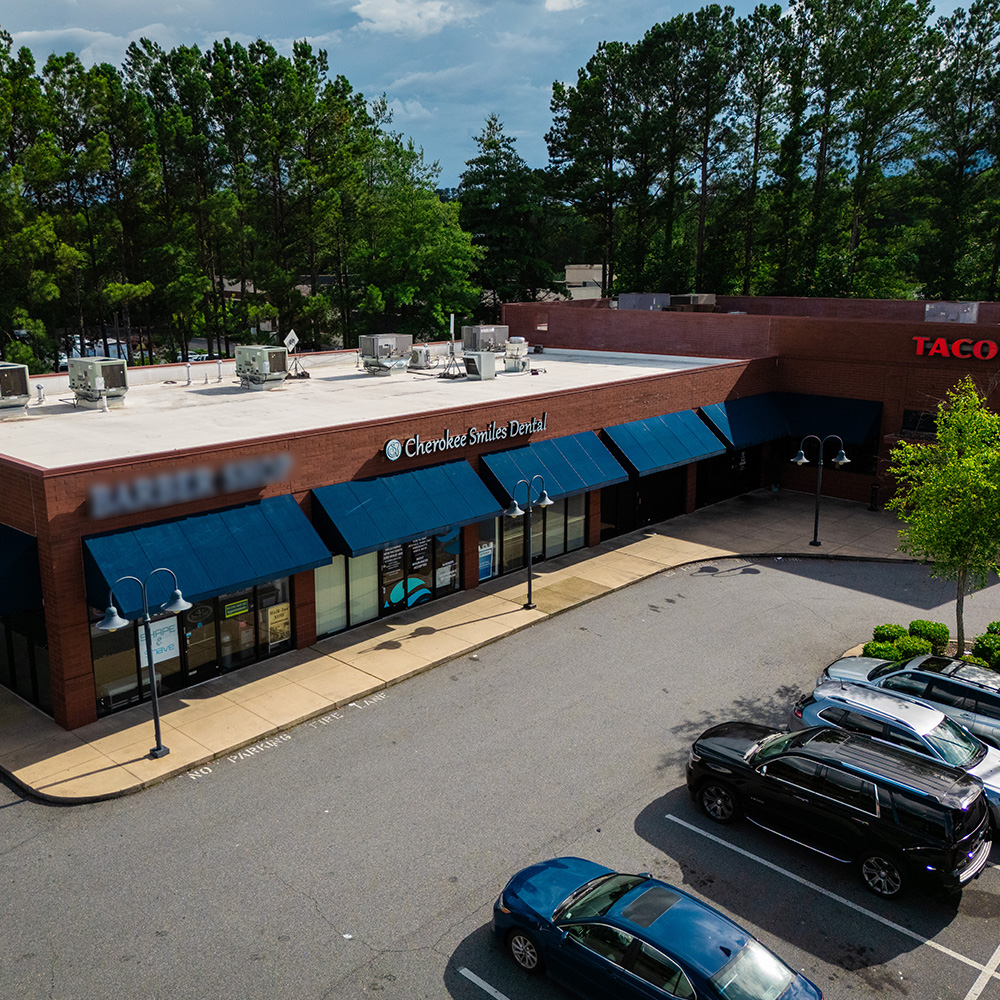 The image shows a storefront with a parking lot, featuring a prominent building sign, multiple parked cars, and a clear sky above.