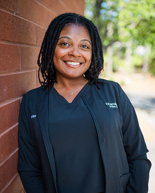 The image shows a woman smiling at the camera, wearing a black jacket with a name tag, standing against a brick wall.