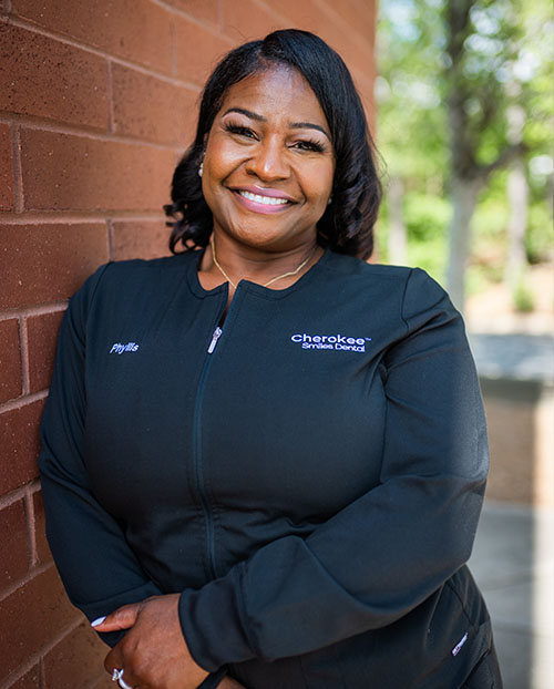 The image features a woman standing against a brick wall with a smile on her face, wearing a black jacket with a logo on the left chest area and a name tag that reads Cherokee.