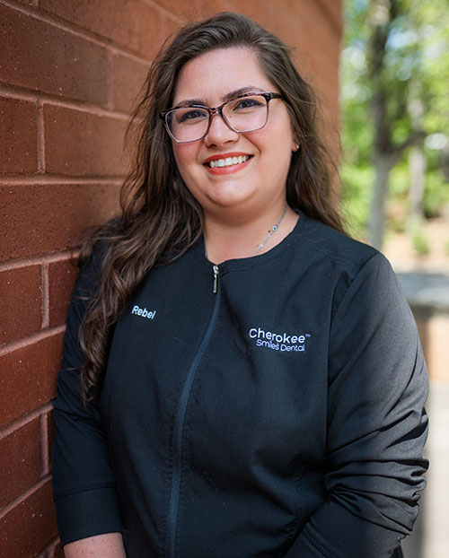 The image shows a woman posing against a brick wall, wearing glasses and a black apron with a logo on it, smiling towards the camera.