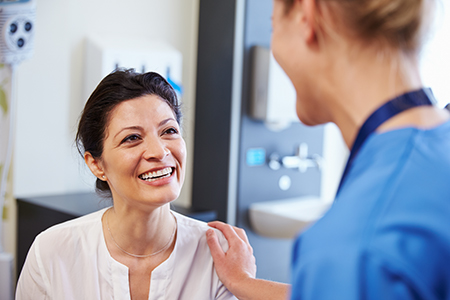 An image featuring two women in a medical setting one appears to be a patient receiving care from a healthcare professional who seems to be a nurse or doctor, with both smiling and engaged in conversation.