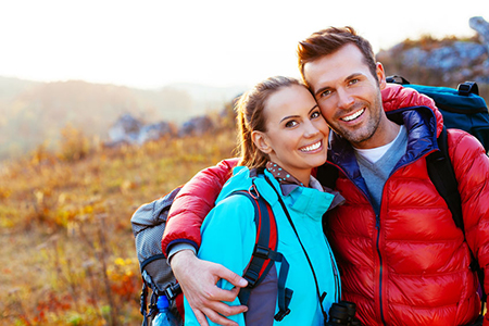 A man and woman are smiling and hugging each other outdoors, with the man wearing a backpack and both dressed for cold weather activities.
