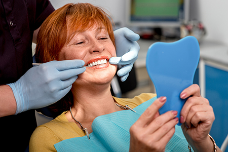 A woman is sitting in a dental chair with a blue mask on her face, smiling at the camera while a dentist looks at her teeth.