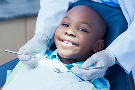 A young boy sitting in a dental chair with a smiling expression, receiving dental care from a professional wearing protective gloves and a mask.