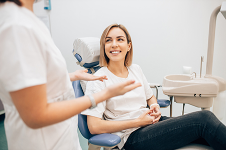 The image shows a woman seated on a dental chair with her eyes closed, smiling slightly, while being attended to by a dental professional in a clinical setting.