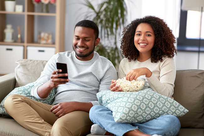 A man and woman sharing a moment on a couch, enjoying popcorn while watching something on a smartphone.